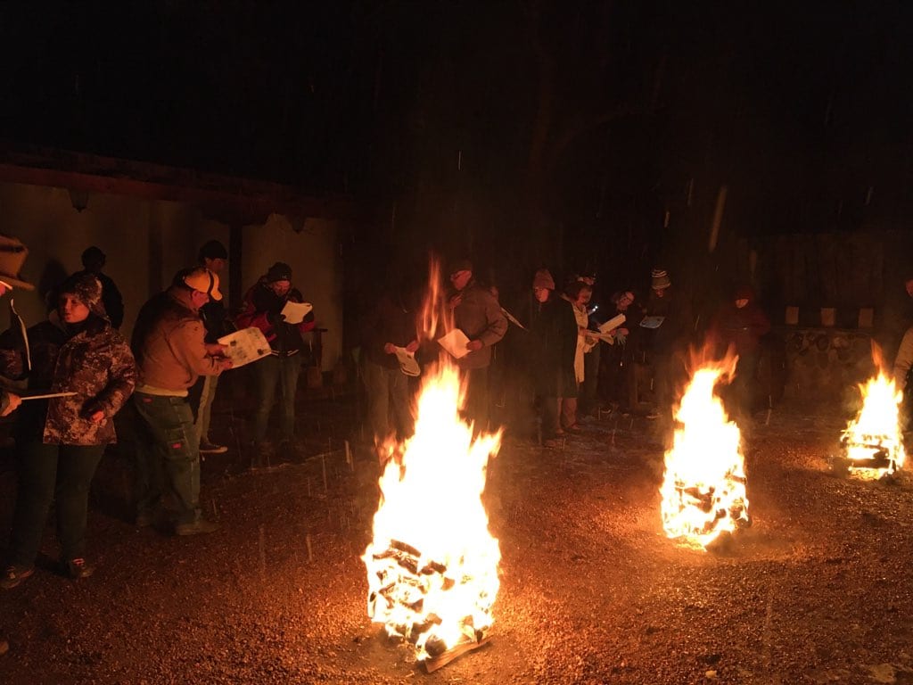 People singing by piñon wood bonfires on a Santa Fe street on Christmas Eve, with an adobe wall behind them